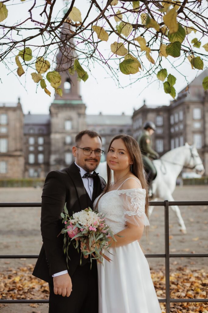 Heiraten in Dänemark: Ein Brautpaar posiert vor Christiansborg Slot mit einem Reiter auf einem weißen Pferd im Hintergrund.